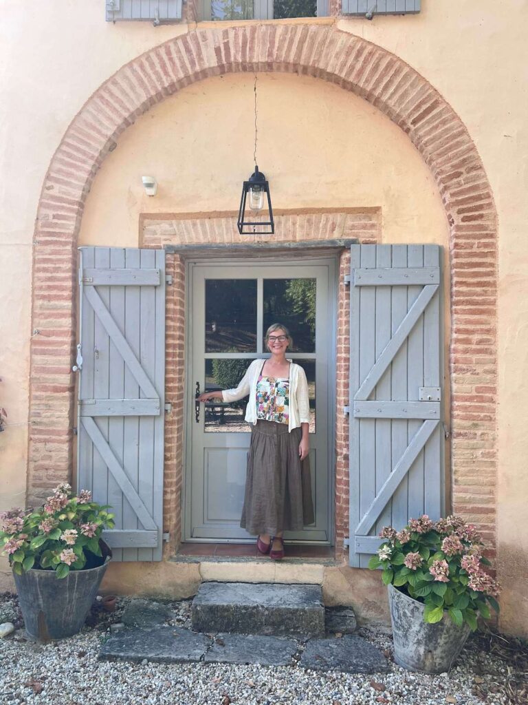 Kelly Rae Roberts standing joyfully in a French countryside doorway.